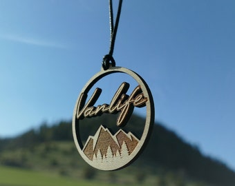 May include: A wooden car air freshener with the word "Vanlife" and a mountain scene. The air freshener is hanging from a rearview mirror.