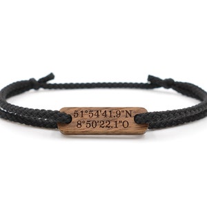 Könnte beinhalten: Schwarzes Seilarmband mit einem rechteckigen Mittelstück aus Holz. Das Holz ist mit Koordinaten graviert: 51°54'41.9"N 8°50'22.1"O. Das Armband ist an beiden Enden gebunden.