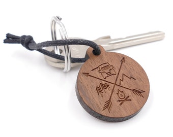 May include: A wooden keychain with a compass design featuring a campfire, mountains, and a van. The keychain is attached to a black cord and a silver key ring.