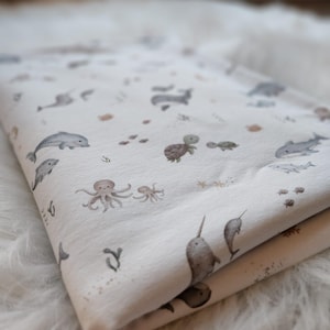 May include: A folded piece of off-white fabric featuring a repeating pattern of watercolor-style sea creatures including dolphins, narwhals, turtles, octopuses, and sharks in muted shades of gray, brown, and tan, scattered with small aquatic plants.