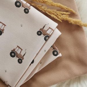 May include: A stack of beige fabric with a tractor pattern, featuring brown tractors with black wheels. The fabric is folded over a solid tan fabric. A dried wheat stalk rests on top of the fabric.