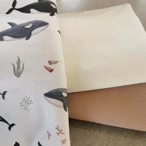 Op de afbeelding: Een stapel stoffen. De hoofdstof heeft een witte achtergrond met illustraties van orka's, zeewier en schelpen. Het is gevouwen over een beige en een lichtbruine stof, waardoor een gelaagd effect ontstaat. De orka's zijn zwart en wit, het zeewier en de schelpen zijn in gedempte tinten.