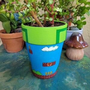 May include: A green, blue, and brown ceramic planter painted with a Super Mario Bros. theme. The planter has a green top, a blue middle section with a white cloud and a question mark block, and a brown bottom with a green bush and three gold coins.