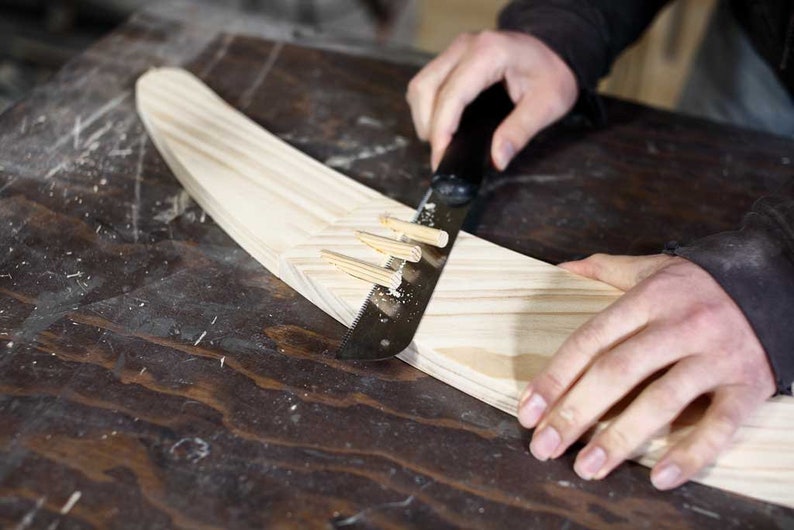 Puede incluir: Una persona utiliza una sierra de mano para cortar una pieza de madera. La madera se mantiene en su lugar mediante varias clavijas de madera.
