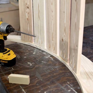 Puede incluir: Un taladro inal&aacute;mbrico amarillo y negro se est&aacute; utilizando para unir una pieza de madera a una pieza de madera m&aacute;s grande. La madera es de color marr&oacute;n claro y tiene un acabado natural. Una botella de pegamento para madera es visible en el fondo.