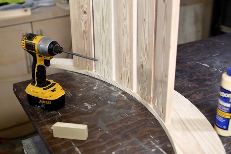 Puede incluir: Un taladro inal&aacute;mbrico amarillo y negro se est&aacute; utilizando para unir una pieza de madera a una pieza de madera m&aacute;s grande. La madera es de color marr&oacute;n claro y tiene un acabado natural. Una botella de pegamento para madera es visible en el fondo.