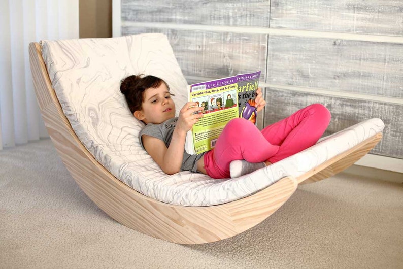 Puede incluir: Un ni&ntilde;o se sienta en una silla mecedora de madera con un coj&iacute;n estampado en blanco y gris. El ni&ntilde;o est&aacute; leyendo un libro titulado "Garfield Eats, Sleeps, and Hates Mondays" con una portada morada y amarilla.