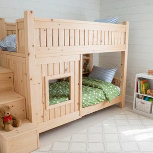 Puede incluir: Una litera de madera clara con una escalera incorporada y una pequeña ventana. La cama tiene una litera superior con una almohada a rayas azules y blancas y una litera inferior con ropa de cama verde. Un oso de peluche está en las escaleras.