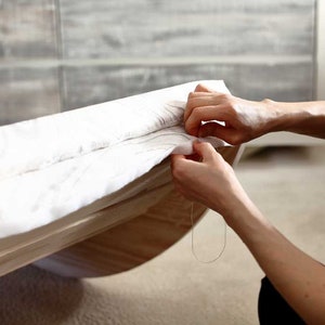 Puede incluir: Una persona cosiendo tela blanca a un marco de madera. La tela se est&aacute; tensando y la persona est&aacute; usando una aguja e hilo para sujetarla en su lugar.