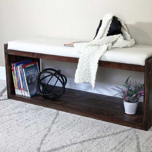 May include: A dark brown wooden bench with a white cushioned top. The bench has a shelf underneath for storage, which is filled with books and a decorative metal orb. A white throw blanket and a black purse are placed on top of the bench.