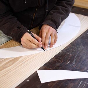 Puede incluir: Una persona trazando una l&iacute;nea curva en un trozo de papel blanco con un l&aacute;piz. El papel est&aacute; sobre una tabla de madera.
