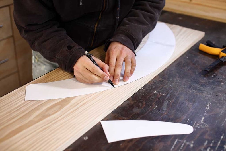 Puede incluir: Una persona trazando una l&iacute;nea curva en un trozo de papel blanco con un l&aacute;piz. El papel est&aacute; sobre una tabla de madera.