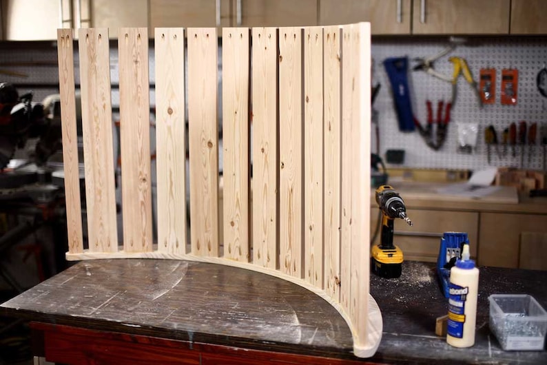 Puede incluir: Un marco de madera curvo hecho de m&uacute;ltiples listones de madera, unidos con tornillos. El marco est&aacute; sentado sobre una mesa de madera, con un taladro, pegamento y un peque&ntilde;o recipiente de tornillos visibles en el fondo.