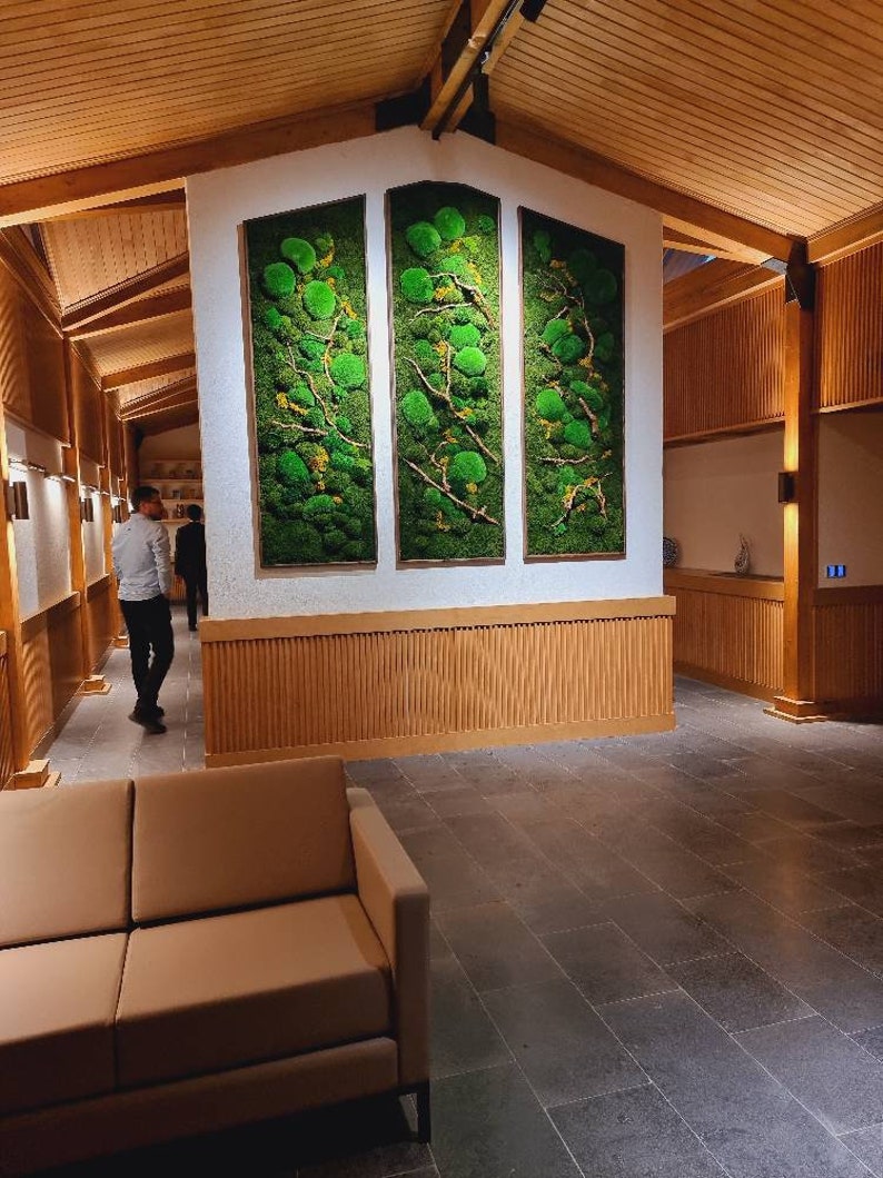 Op de afbeelding: Drie verticale rechthoekige wandkunstwerken met groen mos en takken. De kunst is omlijst in lichtbruin hout en gemonteerd op een witte muur. De kamer heeft een lichtbruin houten plafond en muren, met een bruine bank op de voorgrond.