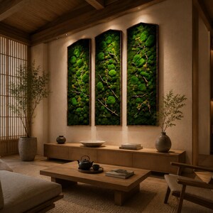 Op de afbeelding: Een moderne woonkamer met een natuurlijke uitstraling. Drie verticaal ingelijste moskunstwerken hangen aan de muur. Een houten salontafel, een bank en een stoel staan in de kamer. Een groot raam met uitzicht op bomen bevindt zich aan de linkerkant.