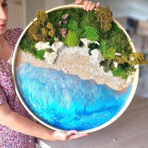 Puede incluir: Una obra de arte mural redonda que representa una escena de playa en miniatura. La obra incluye una playa arenosa, rocas blancas, musgo verde y un océano de resina azul, todo dentro de un aro de bordado de madera.