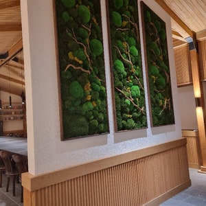 Op de afbeelding: Drie verticale wandkunstwerken met bewaard groen mos en takken in houten frames. De moskunst voegt een natuurlijk, gestructureerd element toe aan het interieur. De frames zijn warmbruin en complementeren het groene mos.
