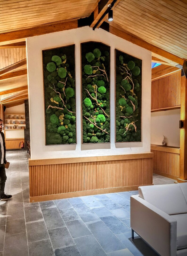 Op de afbeelding: Drie ingelijste verticale wandkunstwerken met groen mos en takken. De moskunst is geplaatst tegen een witte muur met een houten rand. De kamer heeft een modern design met houten balken en een grijze tegelvloer.