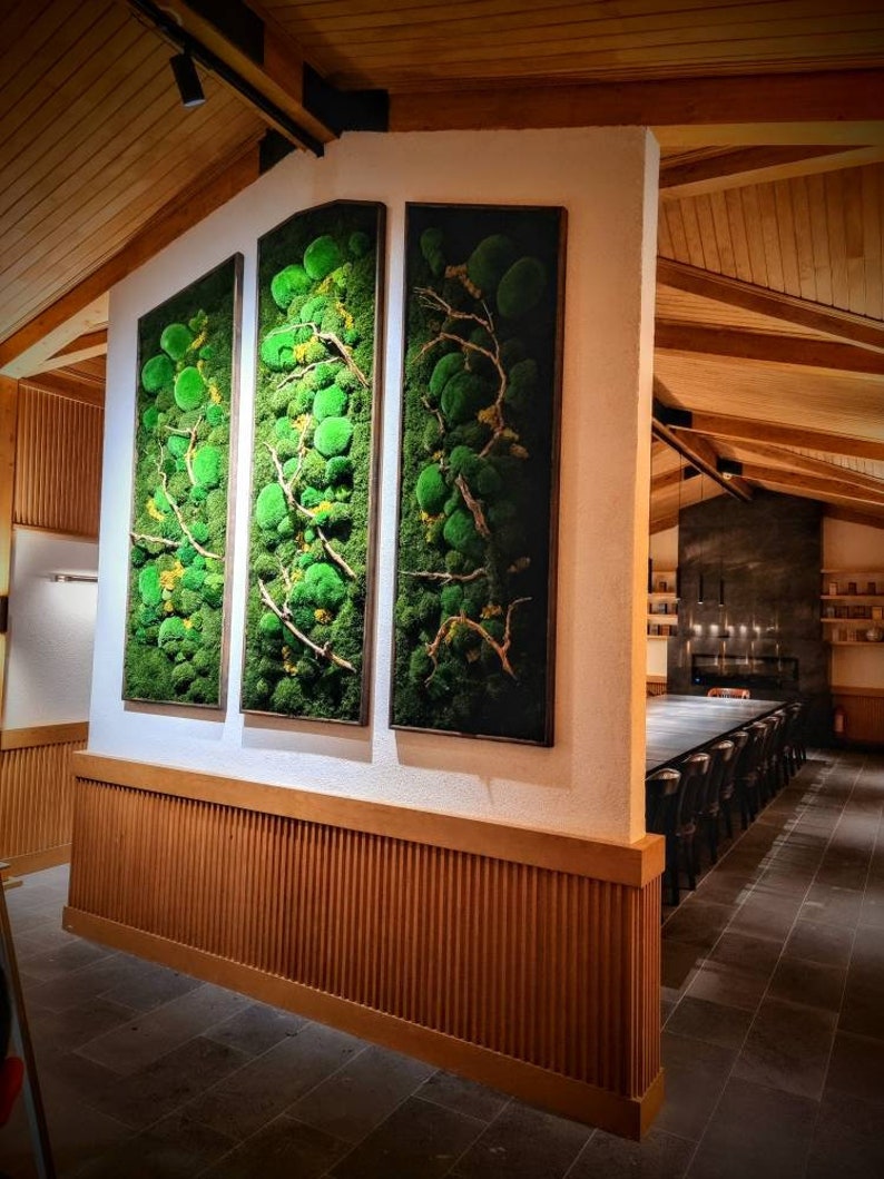 Op de afbeelding: Drie ingelijste verticale wandkunstwerken met levendig groen mos en takken. De kunstwerken worden tentoongesteld op een witte muur met een houten rand. De kamer heeft een rustieke, natuurlijke uitstraling met houten balken en een lange tafel.