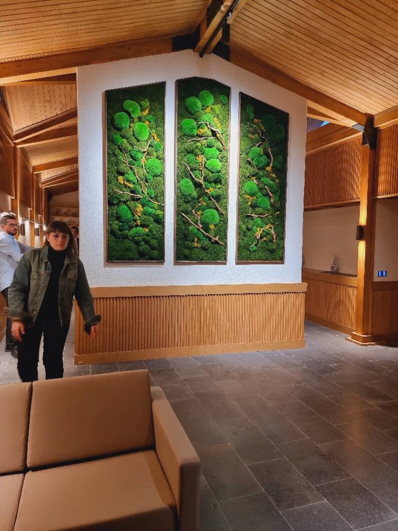 Op de afbeelding: Drie verticale rechthoekige wandkunstwerken met een groen mosontwerp en houten frames. Het mos heeft een mix van groene en gele tinten, met dunne takken. De kunst is gemonteerd op een witte muur boven een houten paneel. De kamer heeft een houten plafond en een betegelde vloer.