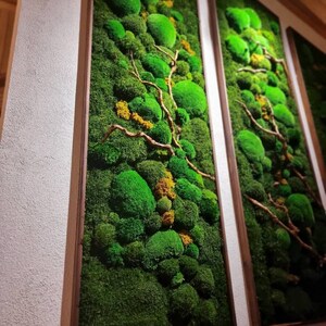 Op de afbeelding: Drie verticale wandkunstwerken met levendig groen mos en kleine gele accenten. Het mos is gerangschikt in een natuurlijk, gestructureerd patroon, met houten takken. De frames hebben een warme bruine kleur.