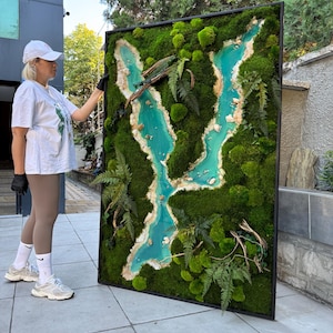 Puede incluir: Una gran obra de arte mural rectangular con un marco negro, que muestra un paisaje de musgo verde con un diseño de río turquesa. La obra de arte incorpora helechos y elementos naturales, creando una estética única y natural.