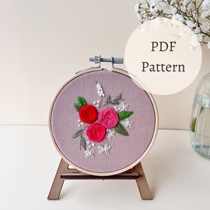 Pode incluir: Um padrão PDF para bordado com um design floral com três rosas cor-de-rosa e vermelhas e flores brancas em um tecido roxo claro em um aro de bordado de madeira.