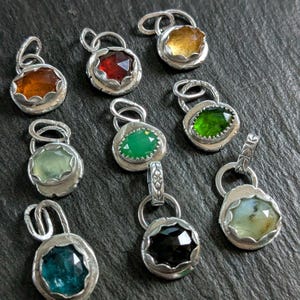 May include: A collection of nine silver pendants, each featuring a faceted gemstone in various colours including amber, red, yellow, light green, dark green, black, and blue, with a scalloped metal setting and a small circular bail for hanging.