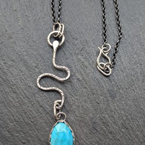 Può includere: Una collana color argento con un ciondolo a forma di serpente e una pietra turchese a forma di goccia. La collana ha una catena scura e un'esclusiva chiusura. La pietra turchese è incastonata in una lunetta d'argento.
