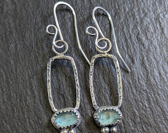 May include: A pair of silver dangle earrings with a light blue gemstone in each. The earrings have a textured rectangular frame and a cluster of three silver beads below the gemstone.