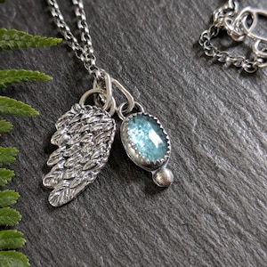 May include: A silver necklace featuring two charms suspended from a delicate chain. One charm is a textured silver wing, and the other is an oval, faceted light blue gemstone set in a silver bezel with a small silver sphere at the base.