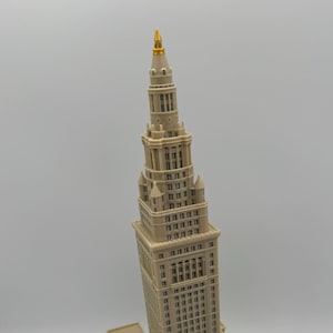 May include: A beige architectural model of a skyscraper with a gold spire. The building has multiple levels, windows, and detailed architectural features. The model is a miniature replica of a famous building.