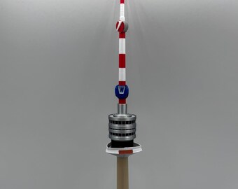 May include: A model of a tower with a beige base and pole, topped with a silver, black, and blue structure. The tower's upper section features red and white stripes and a red tip.