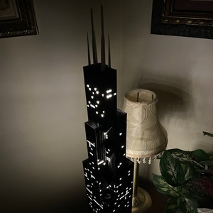 Op de afbeelding: Een zwart Lego-model van de Willis Tower, ook bekend als de Sears Tower, met lampjes die de ramen verlichten. Het model staat op een donker oppervlak met een lamp en een plant op de achtergrond.