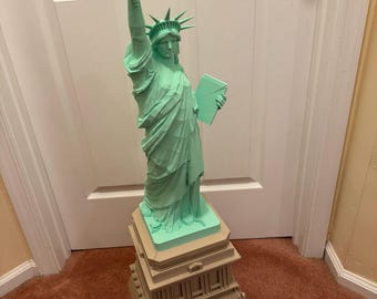 May include: A mint green 3D printed statue of the Statue of Liberty featuring a faceted, geometric design. The figure holds a torch aloft and a tablet in the other hand, standing atop a multi-tiered, tan-colored square pedestal base.
