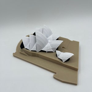 May include: A white 3D printed model of the Sydney Opera House on a brown base.