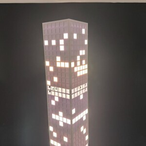 May include: A tall, rectangular lamp resembling a skyscraper. The lamp has a purple and white color scheme, with illuminated white squares mimicking windows. The design evokes a cityscape theme, perfect for home decor.