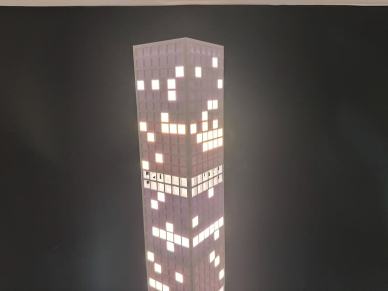 May include: A tall, rectangular lamp resembling a skyscraper. The lamp has a purple and white color scheme, with illuminated white squares mimicking windows. The design evokes a cityscape theme, perfect for home decor.