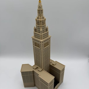 May include: A detailed model of a skyscraper with a tan exterior and a gold-colored spire. The building has multiple levels and architectural details, including windows and decorative elements. The model is a representation of a famous building.