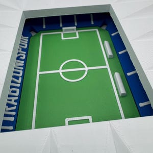 May include: A miniature stadium model with a green field, white markings, and blue seating areas. The stadium's exterior is white, and the text "UNISPOR" is visible in blue. A detailed, three-dimensional sports-themed item.
