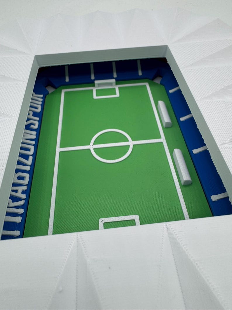 May include: A miniature stadium model with a green field, white markings, and blue seating areas. The stadium's exterior is white, and the text "UNISPOR" is visible in blue. A detailed, three-dimensional sports-themed item.