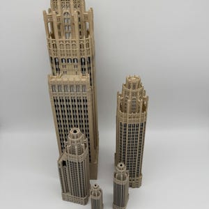 May include: Four beige 3D-printed architectural skyscraper models of varying heights, featuring intricate gothic-style facade details and window patterns. A wooden ruler measuring 12 inches is placed horizontally at the base for scale.
