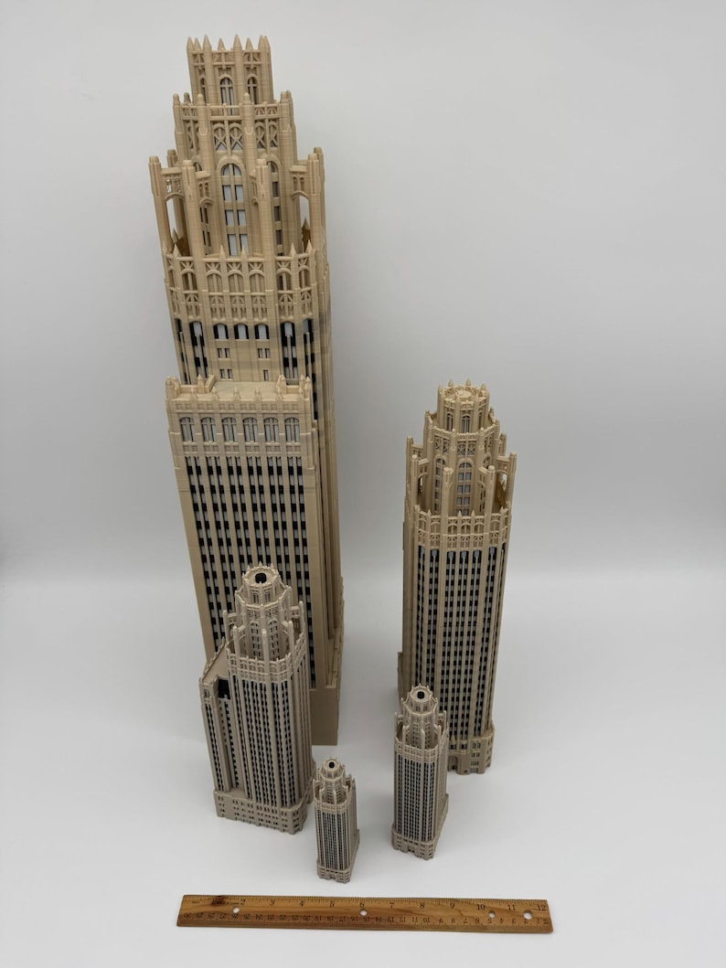May include: Four beige 3D-printed architectural skyscraper models of varying heights, featuring intricate gothic-style facade details and window patterns. A wooden ruler measuring 12 inches is placed horizontally at the base for scale.
