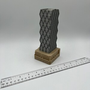 May include: A 3D printed model of a modern skyscraper with a geometric facade. The building is grey and has a brown base. The model is approximately 10 cm tall.