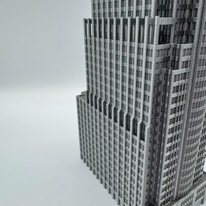 May include: A gray, 3D-printed model of a skyscraper, showcasing intricate architectural details. The building features numerous windows and vertical lines, creating a textured facade. The model is set against a plain white background.