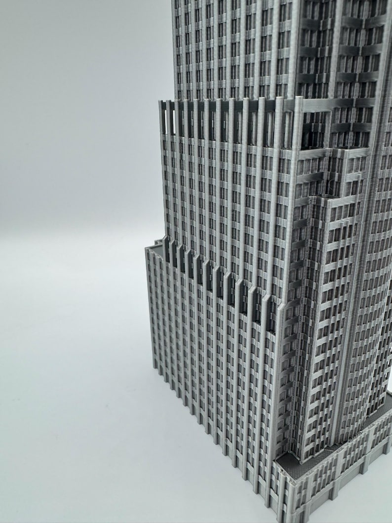 May include: A gray, 3D-printed model of a skyscraper, showcasing intricate architectural details. The building features numerous windows and vertical lines, creating a textured facade. The model is set against a plain white background.