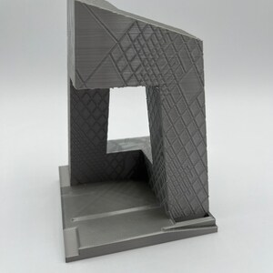 Peut inclure: Mod&egrave;le 3D imprim&eacute; en gris d'un b&acirc;timent avec un design g&eacute;om&eacute;trique. Le b&acirc;timent a une base carr&eacute;e et une haute tour rectangulaire avec une grande ouverture &agrave; l'avant. La tour est surmont&eacute;e d'un toit inclin&eacute;.