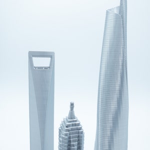 May include: Three silver 3D printed models of skyscrapers. The tallest building is curved and has a modern design. The other two buildings are more traditional in style.
