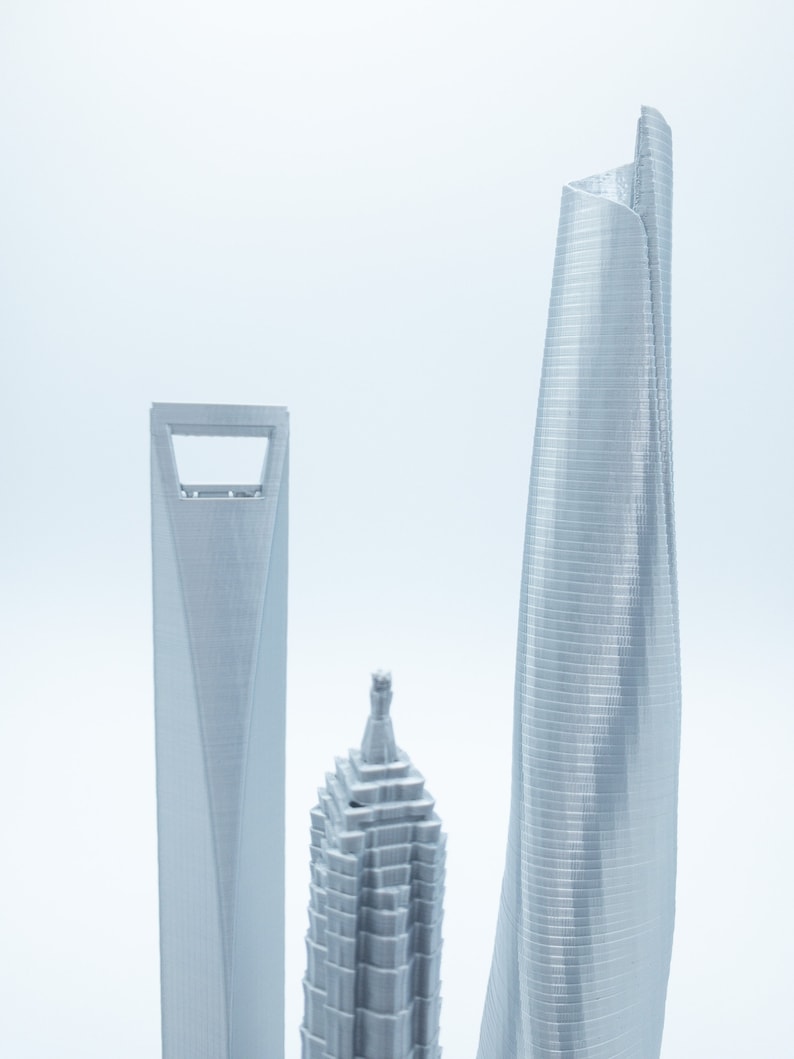 May include: Three silver 3D printed models of skyscrapers. The tallest building is curved and has a modern design. The other two buildings are more traditional in style.
