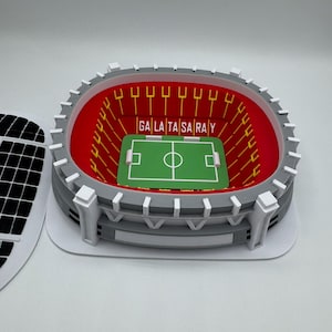 May include: A miniature stadium model with a green field and the text "GALA TA SARAY" in the center. The stadium has red seating, gray walls, and white accents. The model is a detailed representation of a soccer stadium.